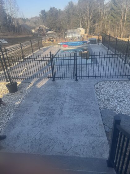 Terrasse en béton estampé gris entourant une piscine creusée, délimitée par une clôture d'aluminium noire